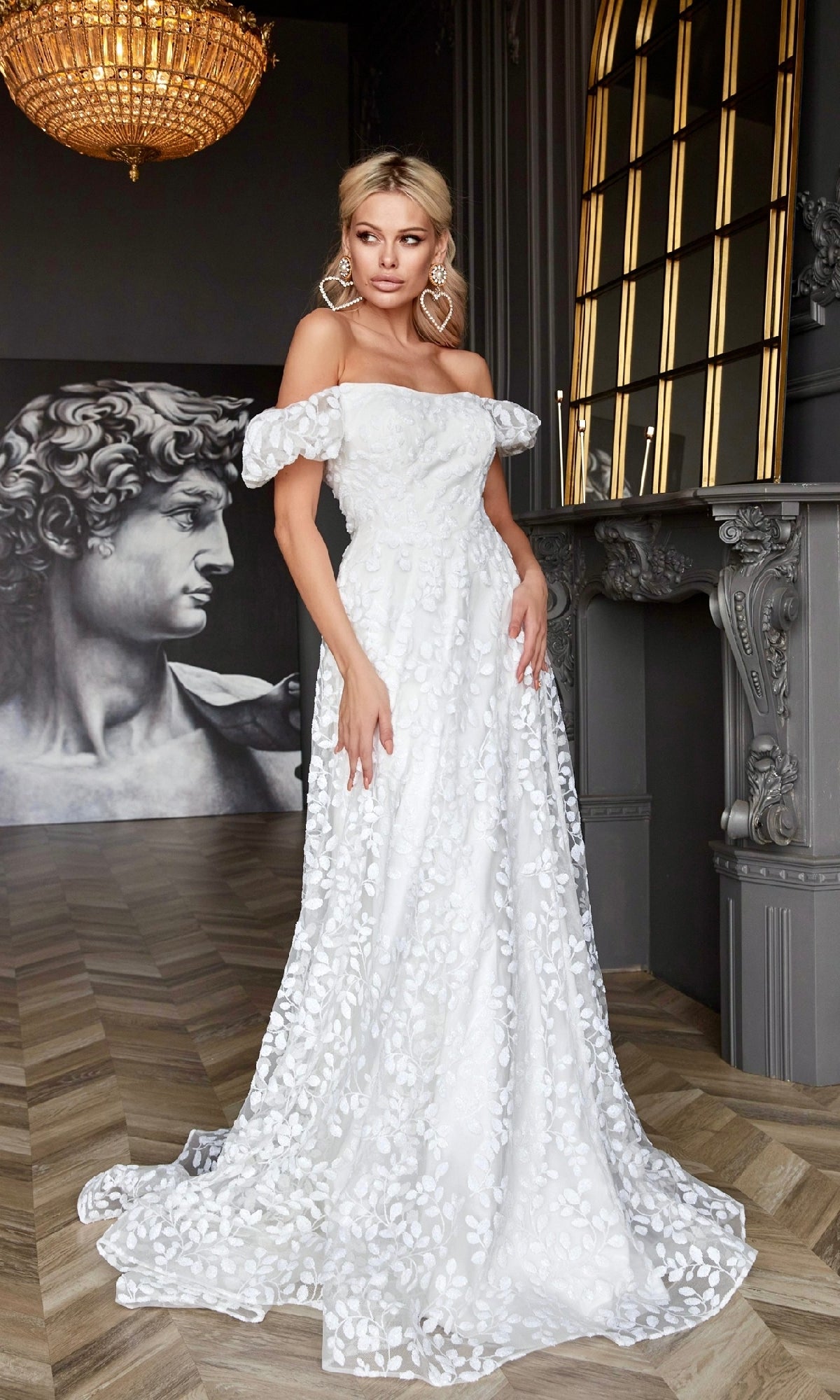 A woman stands indoors beside a gray fireplace, wearing the Tina Holly TK023W long white lace formal dress with floral patterns; a statue and chandelier are seen in the background.